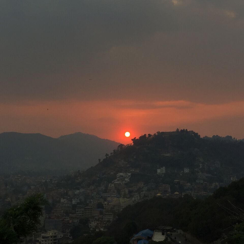 Sunset in Kathmandu - the sun is a red orb sinking over a hilly cityscape.