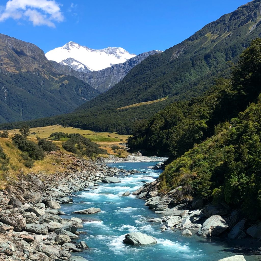 Mt Aspiring National Park in South Island. A stream of blue water cascading over rocks, with a forest going up the slopes behind and snow covered mountains in the distance.