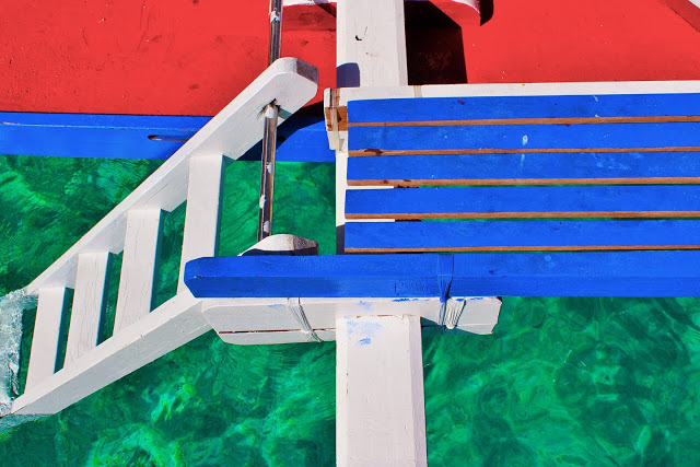 Bright coloured boat decking in red with blue plants and white steps leading into a green ocean.