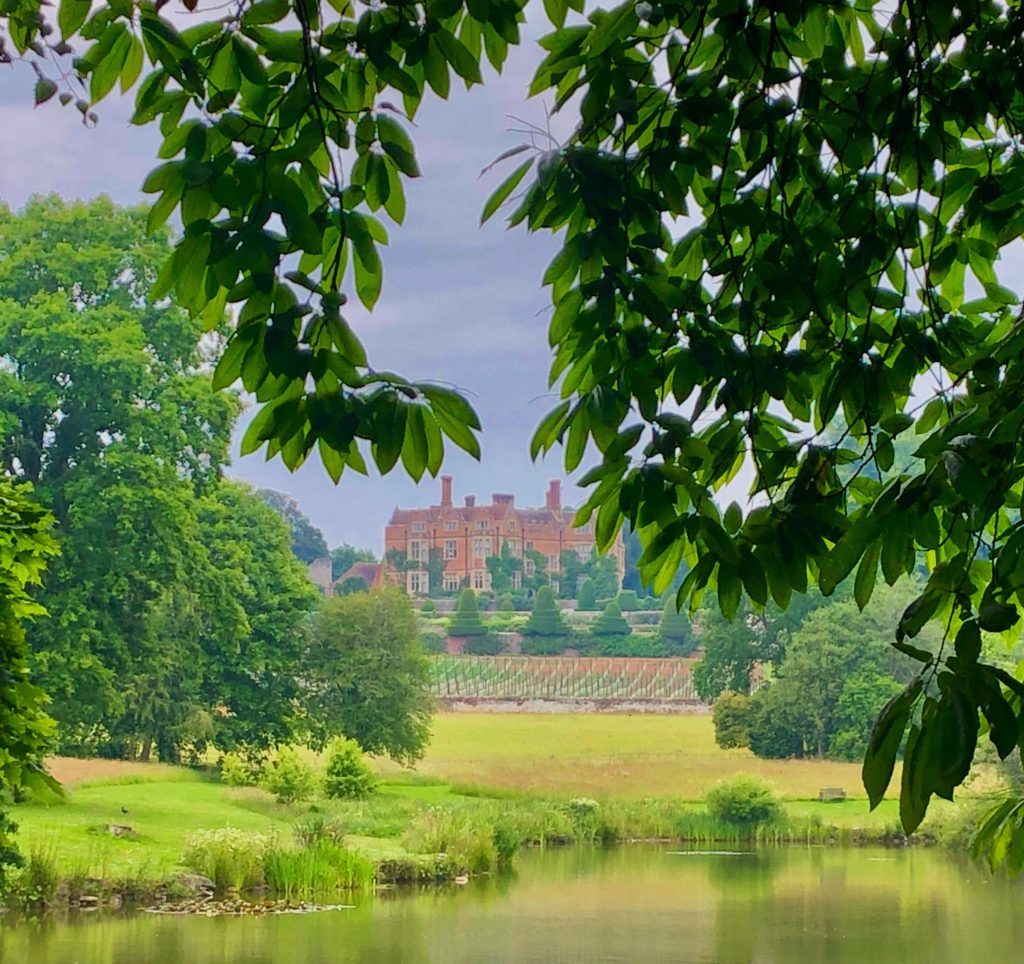 Red brick house - Chilham Castle - visible behind a canopy of green leaves overhanging. There is a lake in front.