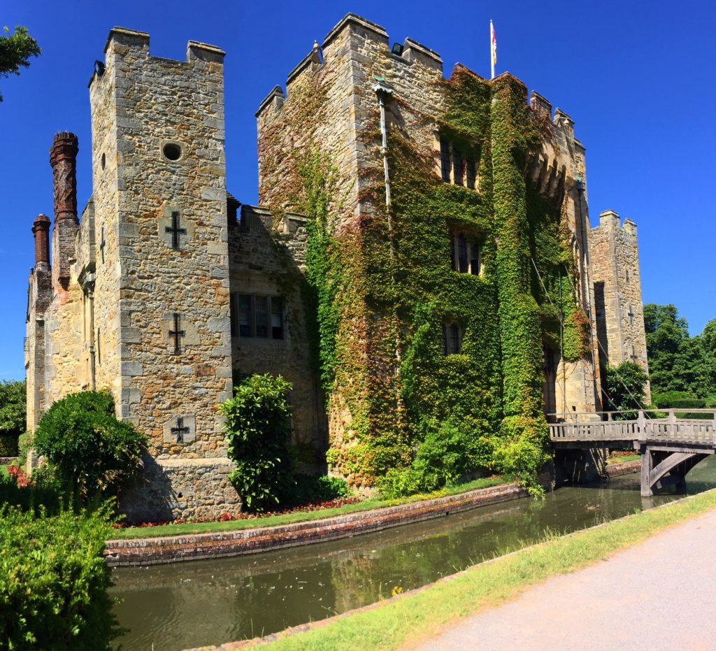 Hever Castle, Kent. The side of Hever Castle covered in ivy with the moat running around and a bridge crossing the water. Blue skies.