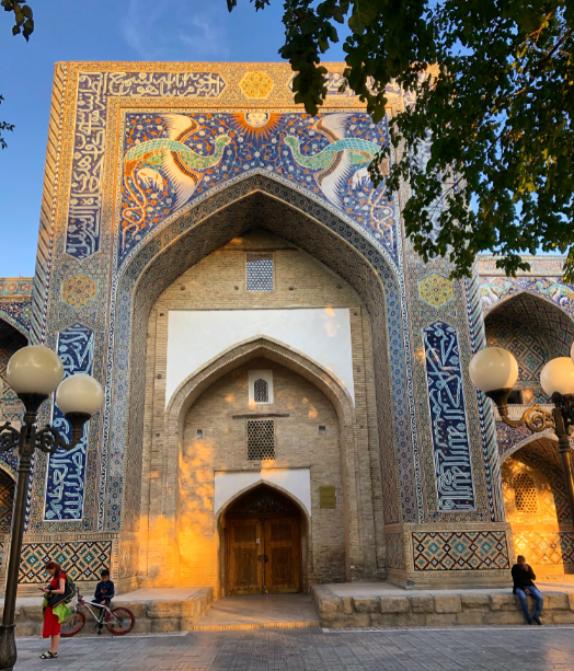 November 2019 - a colourful facade on the front of a madrassa in Bukhara, with people standing out front and golden light from the setting sun.