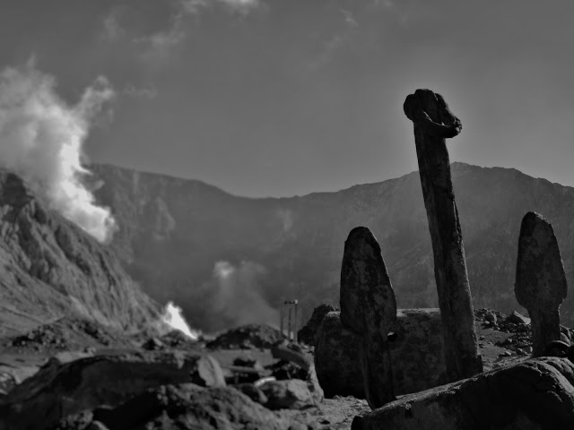 Black and white image showing an anchor in the foreground and steam rising in the background from White Island volcano.