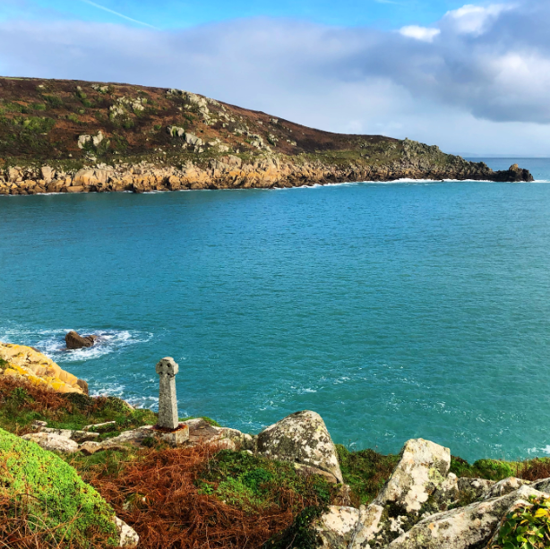 2020. The Cornish coast - a cornflower blue sea, rocky headland and a small stone cross in the foreground, sitting on bracken and rocks.