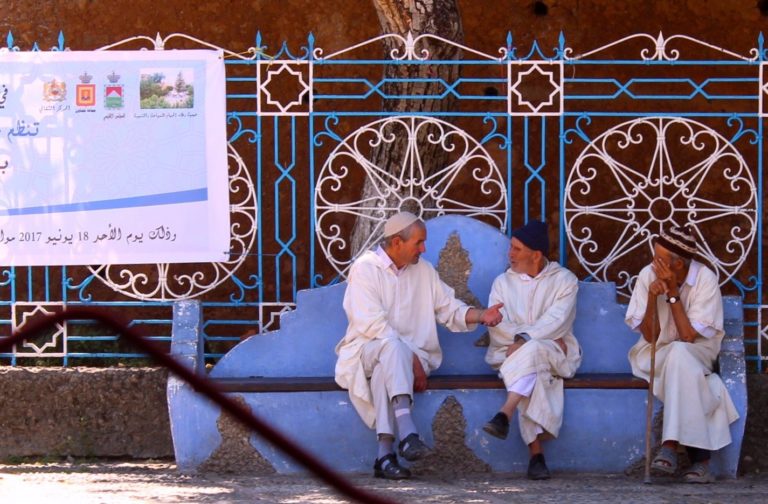travel when not travelling - three men dressed in white robes sitting on a blue bench talking with one another.