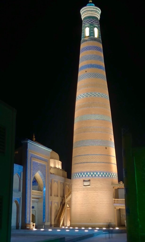 Budget Uzbekistan - a minaret at night time, lit up against a black sky, with a person walking past at the base of the tower.