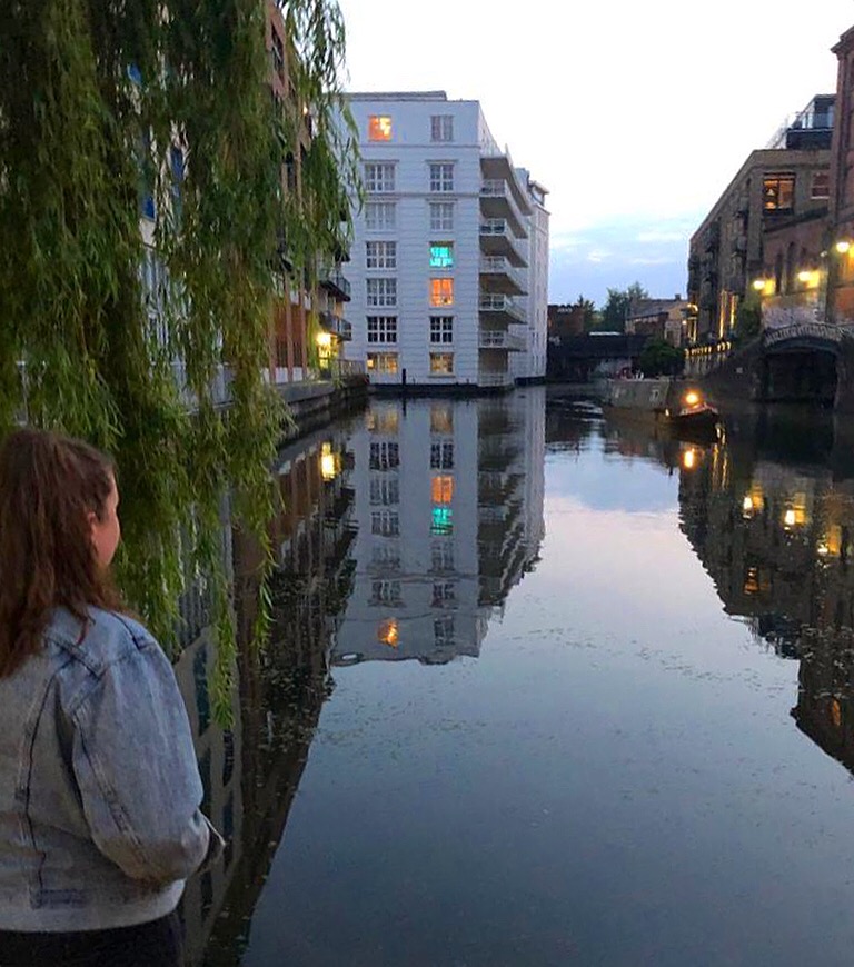 2020. Camden Lock. Still waters reflecting the surrounding buildings, including a white six storey block. I am facing the lock, away from the camera, so only the back of my head can be seen.