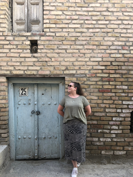 Budget Uzbekistan - a picture of Bethen leaning against a brick wall, wearing a zebra print skirt, a green t-shirt and white trainers. She is smiling, gazing upwards, and standing next to a pale blue small door.