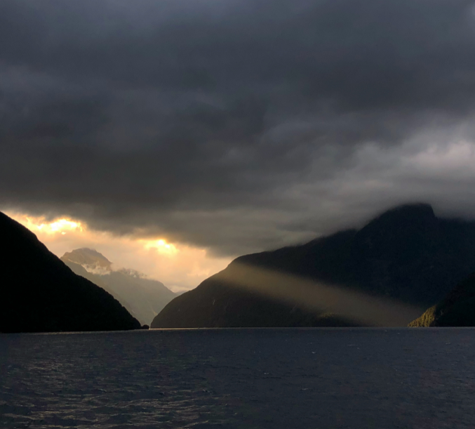 Open water, dark mountains and cliffs and a murky grey sky full of clouds. A ray of light is shining down from the clouds, golden. It hits one cliff on the diagonal.