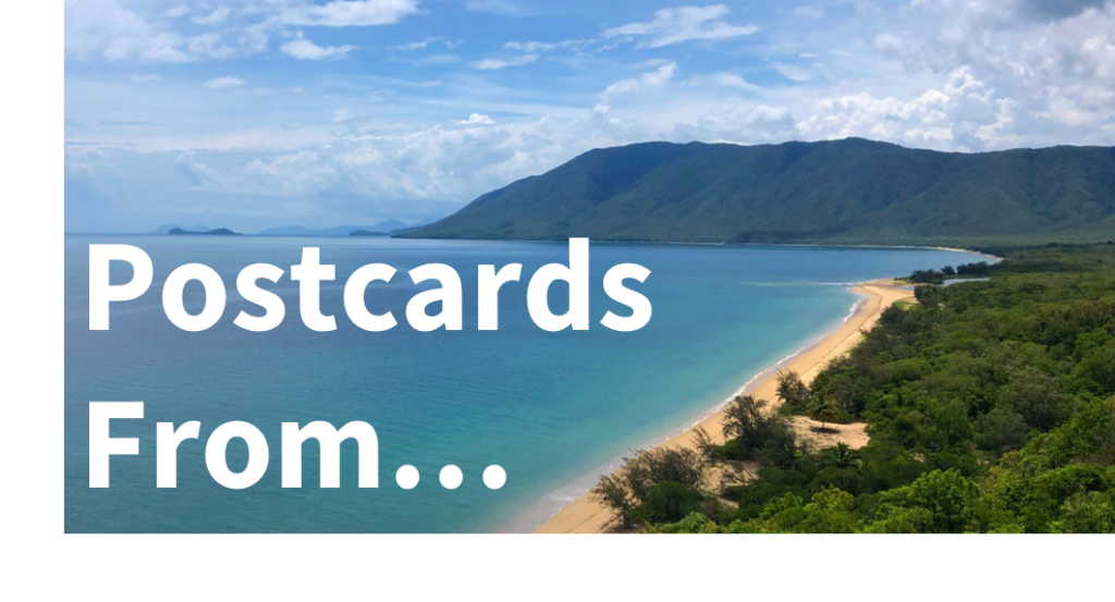 Travel: Postcards From... - text overlaying an image of the blue ocean in Australia, meeting a white sand beach, with trees and a mountain in the background.