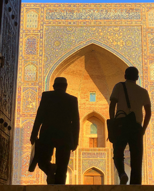 2019 - two men silhouetted against a colourful facade as they exit a doorway. They're both carrying bags and their stances are mirrored - left legs lifted to take their next steps.