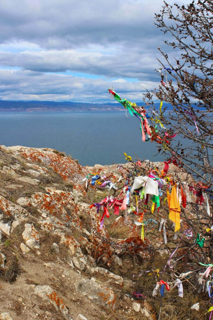 Ribbons and prayer flags tied to a tree on the cliff edge, with Lake Baikal in the background.