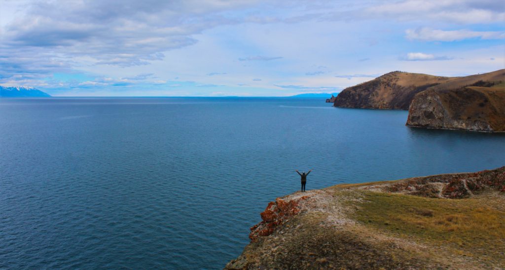 Olkhon Island - Lake Baikal in the background, with cliffs in the foreground and me in the distance standing with my arms in the air.