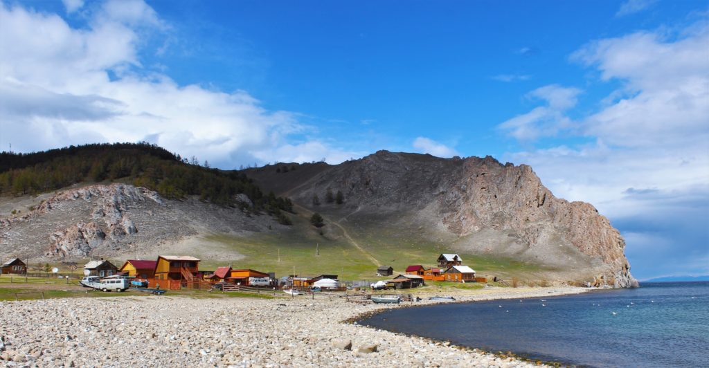 A shingle beach on Olkhon Island, with high cliffs in the background and a group of wooden houses.