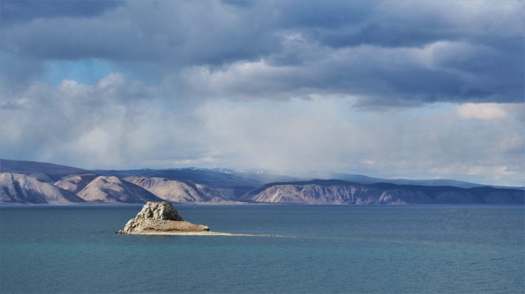 Moody clouds over Lake Baikal. Mountains and cliffs in the distance with a large rock protruding from the lake waters.