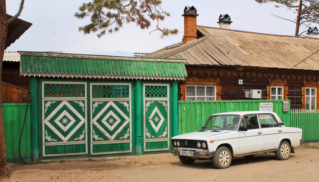 A small white car parked in front of a large green wooden gate on a sandy road. A wooden house is part obscured behind the green fence.