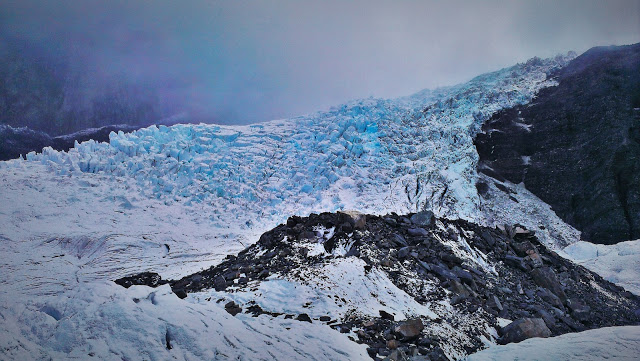 Franz Josef Glacier. Blue ice cascades over a dark rock face, broken, fractured and jagged. There are clouds behind.