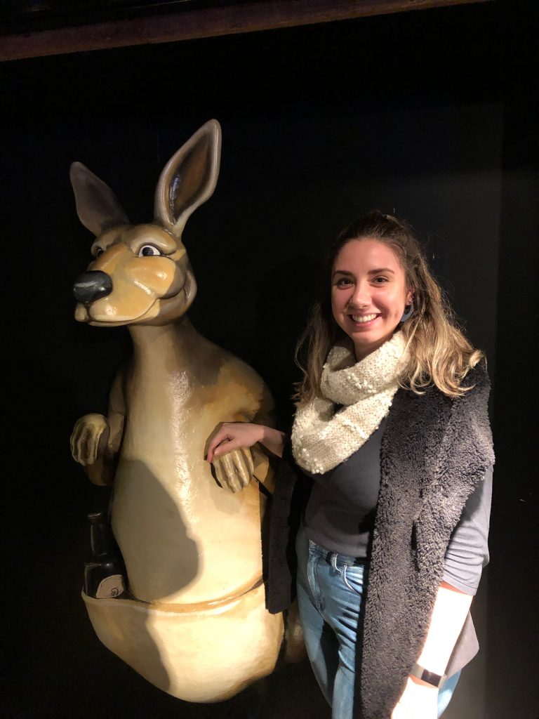 Emma standing next to a statue of a kangaroo at the Guinness Storehouse. She has loose hair, a white scarf and a dark cardigan on. The kangaroo is cartoonish.