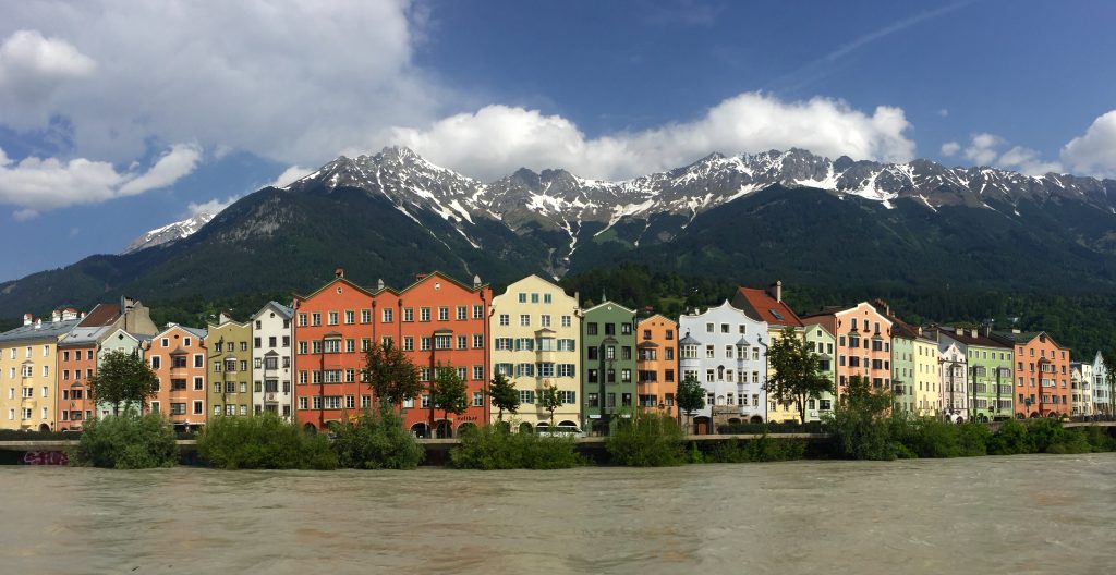 Bucket list - the river Inns flows in the foreground, with a row of colourful houses lining the opposite river bank and snow capped mountains rising up behind.