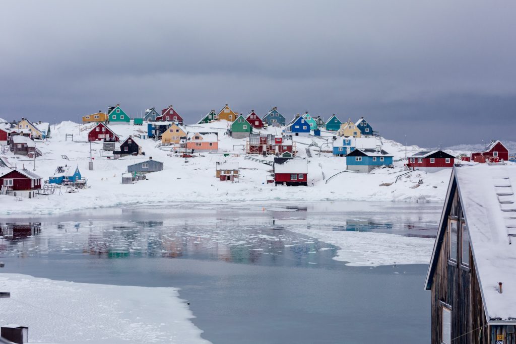 Colourful houses with snow covered roofs on a snowy slope in the background, with ice covered water in the foreground.