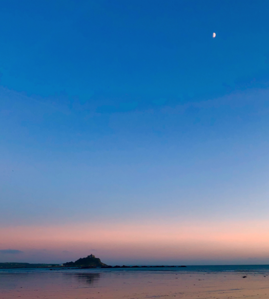 The island of St Michael's Mount is silhouetted against a peach and dark blue sky, in the distance. There is the moon showing in the top right corner and an expanse of beach.