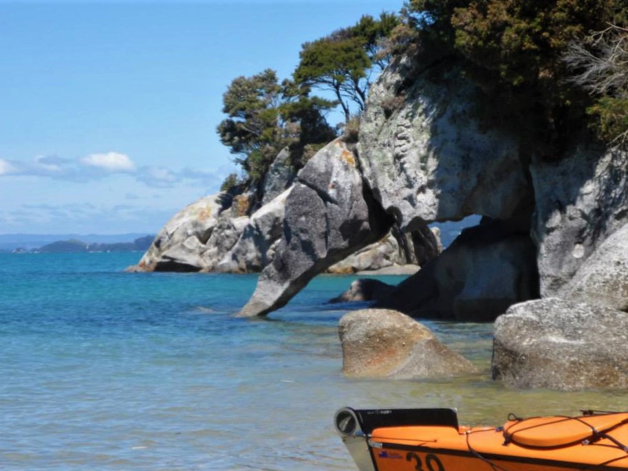 Bucket list - Abel Tasman. An orange kayak in the bottom right of the photo, with rocky cliffs coming down the sea in the background and a blue ocean lapping the shore.