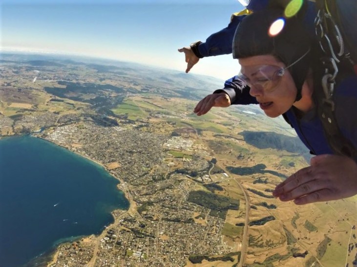 Bucket list. Sky dive over Taupo. Bethen is in the top right of the image, with goggles on. Laid out below is a birds eye view of Taupo township and the lake, blue. The earth is curving behind.