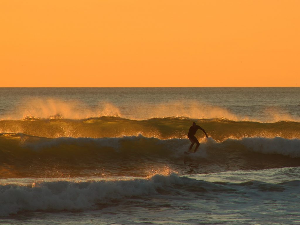 The silhouette of a surfer against three big waves. The sky is orange as it is sunset. Spray is flying off the waves and turning orange in the light.
