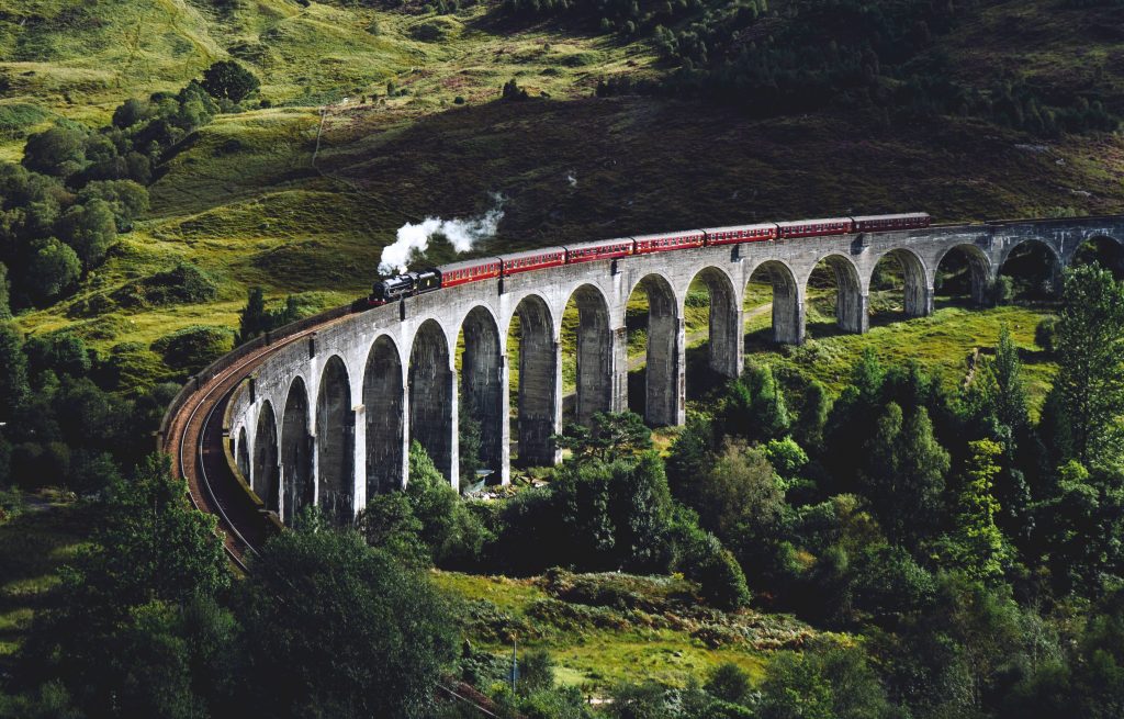 Train travel. A red steam train emitting a column of smoke from its funnel travels the length of a curved viaduct against a backdrop of green hills and trees.