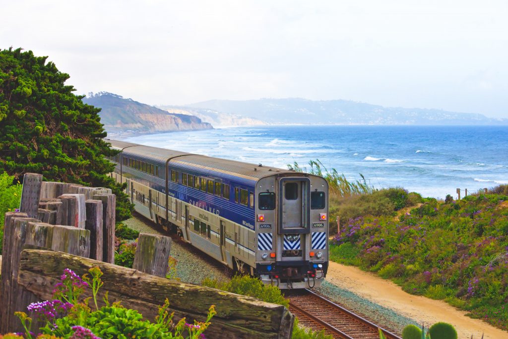 Train travel. A train travelling along a coastline in California. Cliffs and blue sea are in the background, with grasses and flowers along the sides of the train tracks.