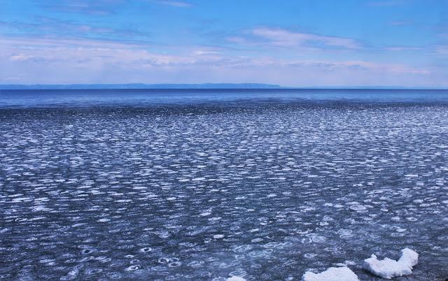 Lake Baikal. The lake is iced over with a thin layer of circle patterned ice, and is very blue. It stretches away into the distance.