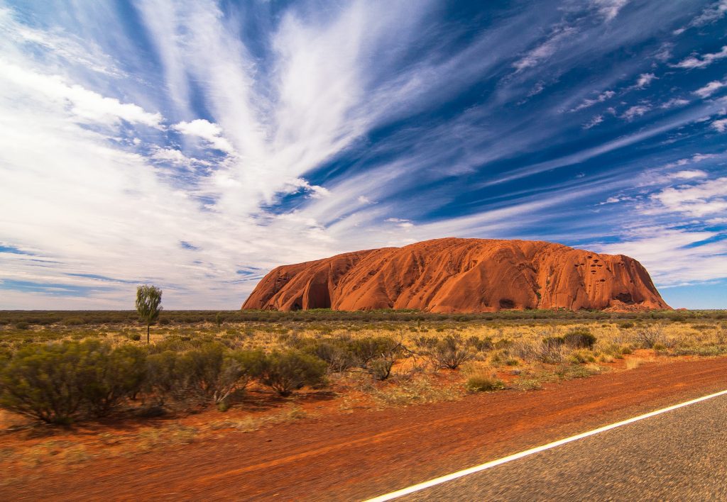 Uluru standing against a backdrop of cloud filled blue sky. The shot is taken from a distance with outback in the foreground.