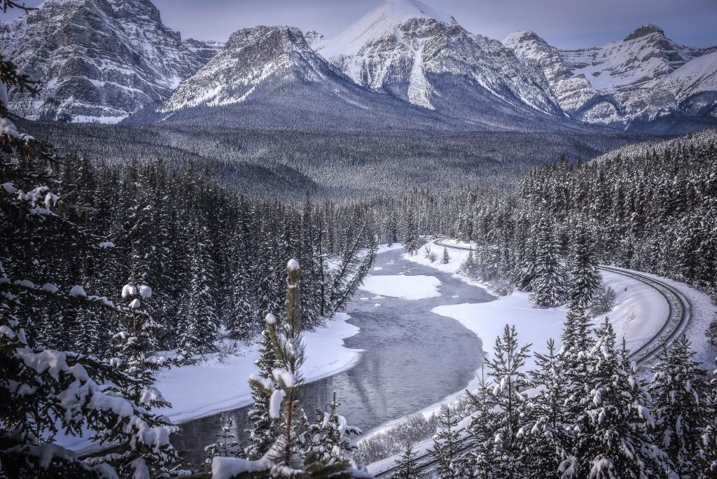 A snow covered landscape with the Canadian Rocky Mountains in the background. In the foreground, a line of train track curves through conifer forests beside an iced up river.