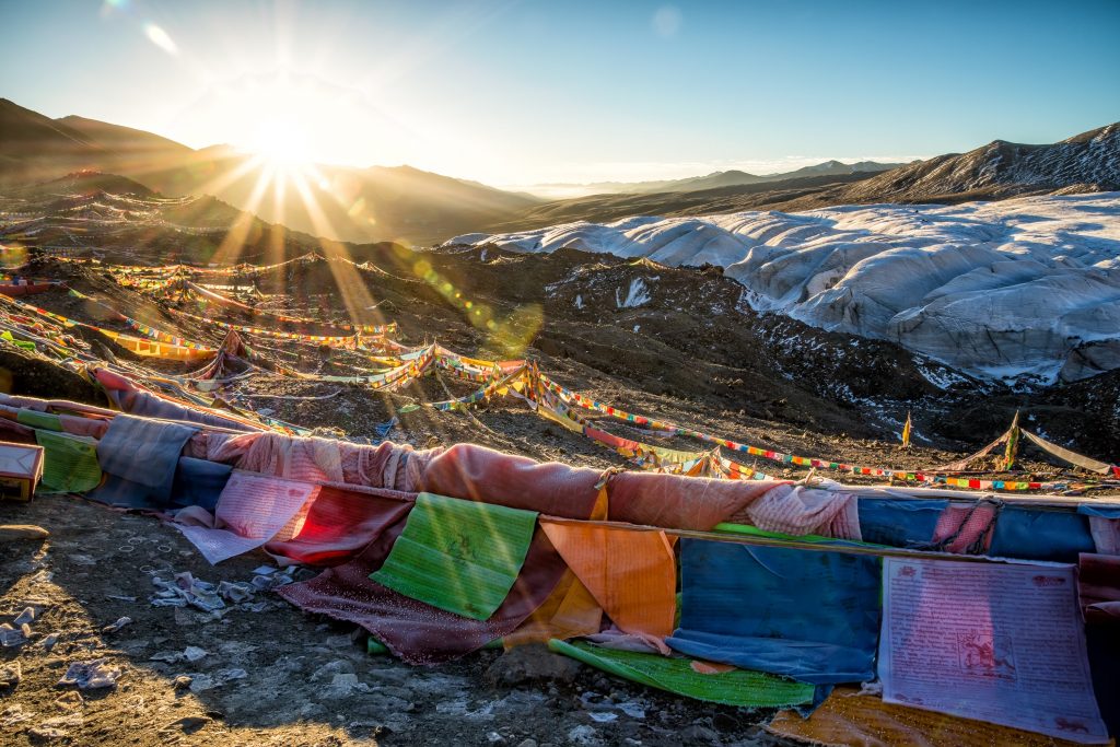 Train travel. Multi coloured prayer flags are strung across rocky ground. The sun is rising behind the scene and shining on a glacier in the background.