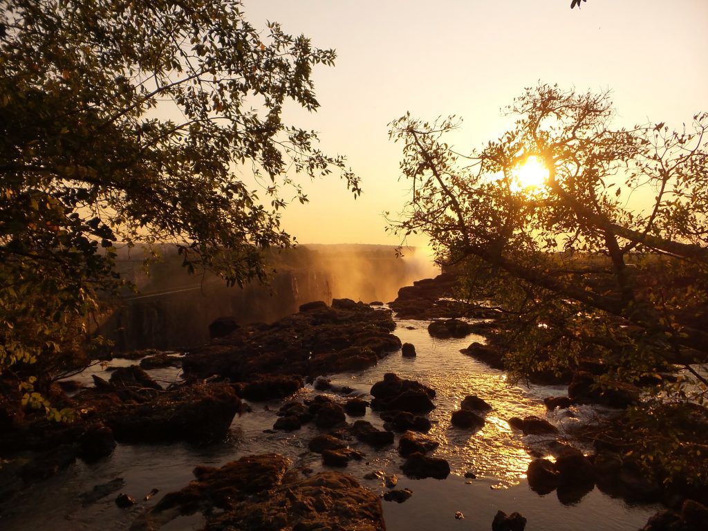 Victoria Falls at sunset. A plume of mist rises up from the waterfalls which was hidden from view. In the foreground, some water flows over dark rocks and there is foliage silhouetted against the orange sky.