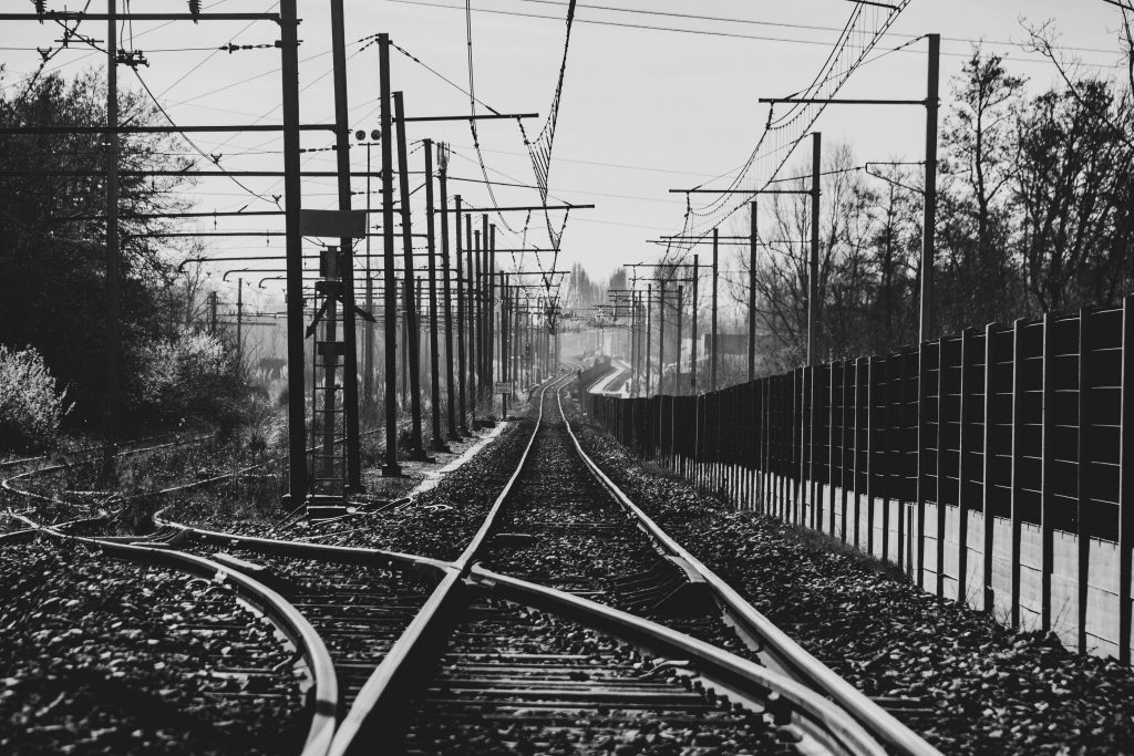 A black and white photo of train tracks leading away into the distance, with overhead power lines and a fence running to the right.