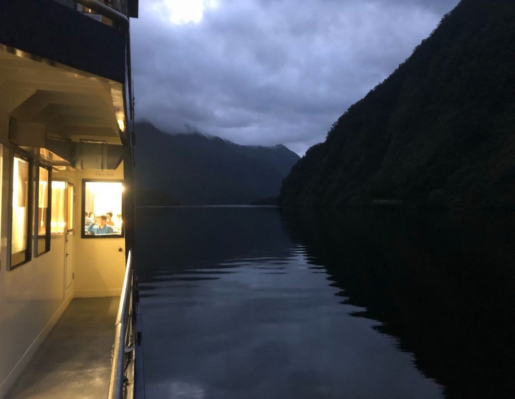 Doubtful Sound. Twilight. The left half of the image shows the lit up deck and people through the windows. The right side shows the dark still water with cliffs coming down the edge.
